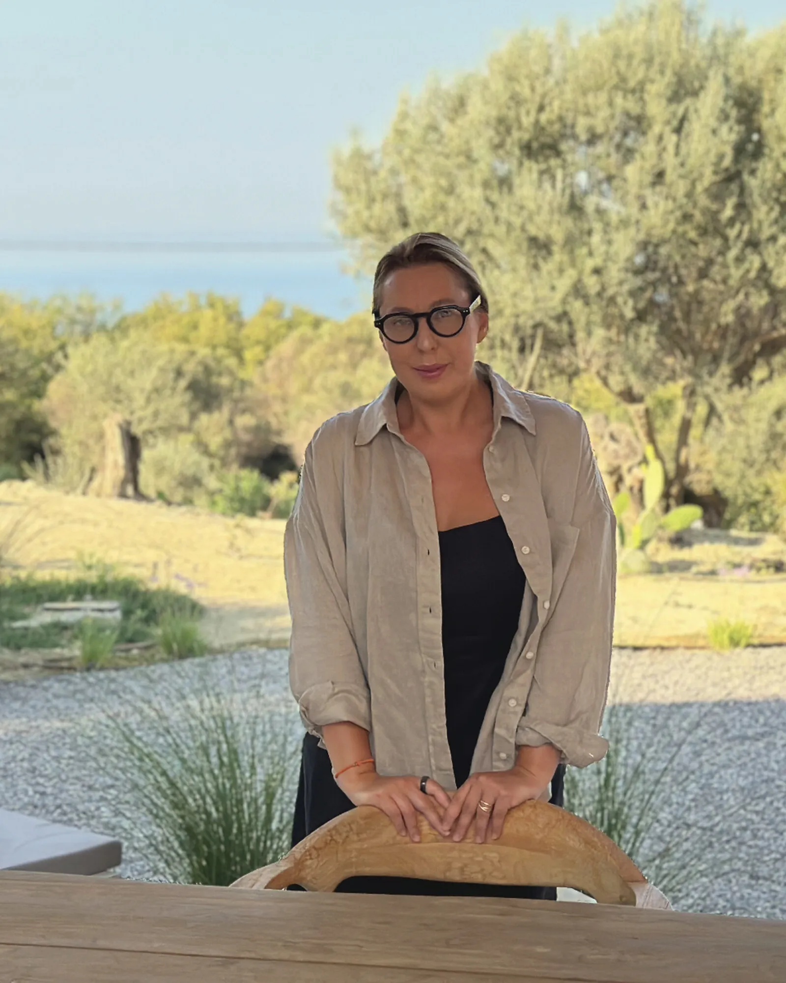 Dr. Olivia Breaban în picioare la o masă de lemn, printre măslini, cu marea în fundal