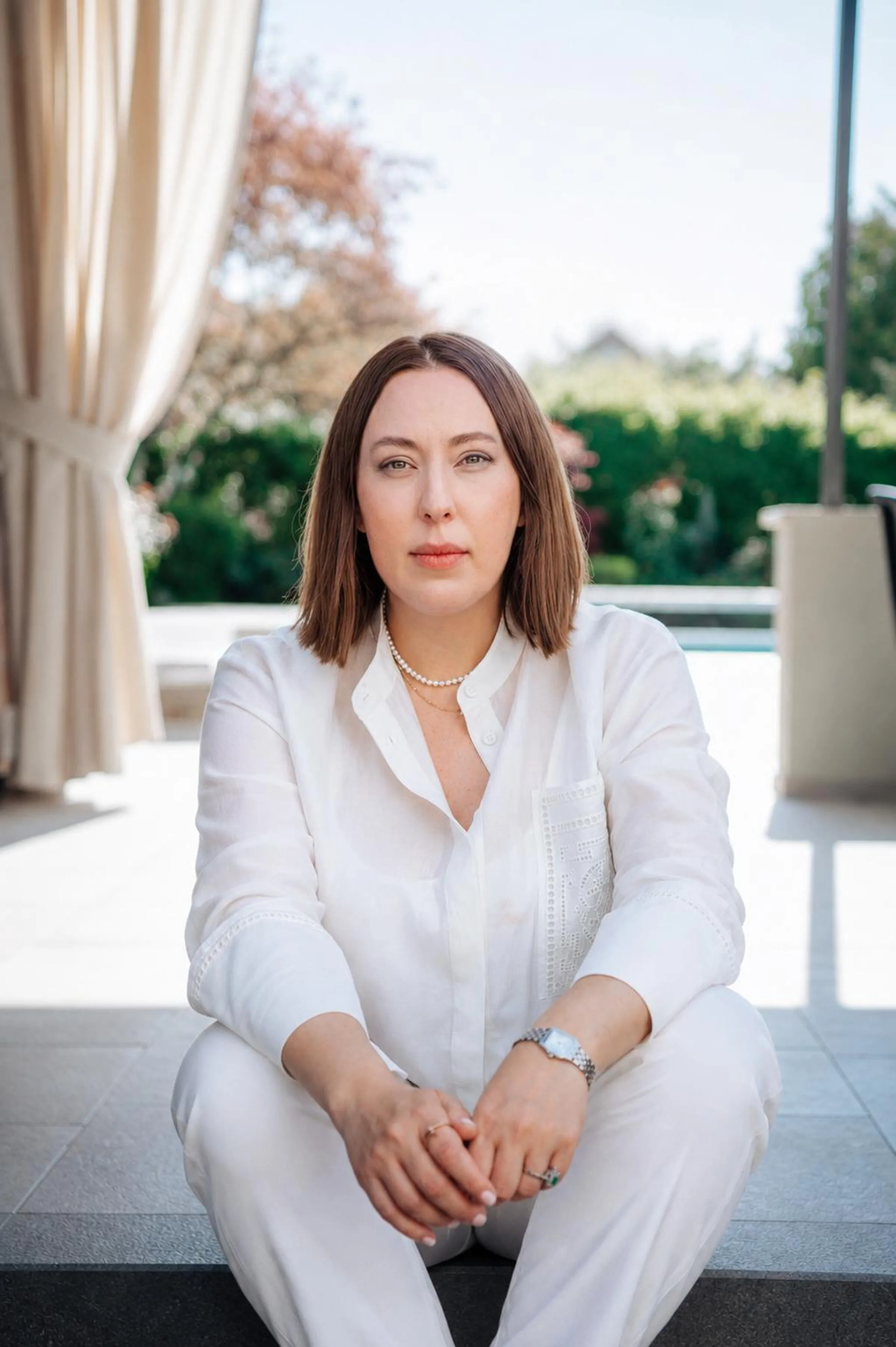 Dr. Olivia Breaban, dressed in white, sitting on a terrace with the garden behind her